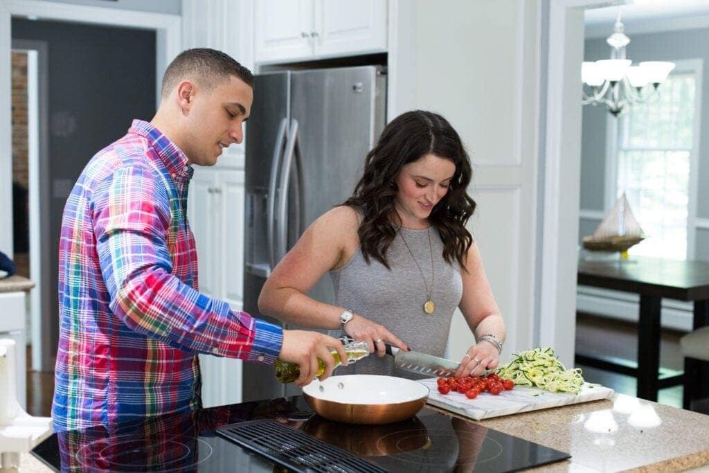 A couple cooking healthy food in a white kitchen. the girl is chopping vegetables and the guy is pouring olive oil into a pan