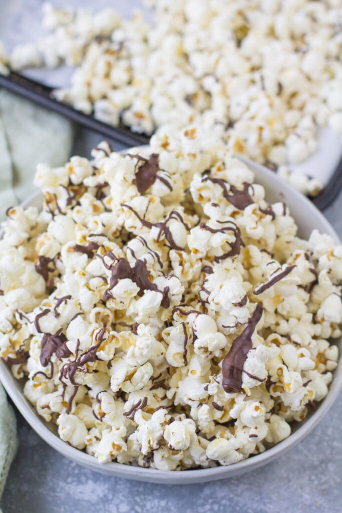 A bowl of chocolate covered popcorn with a tray of chocolate popcorn in the background