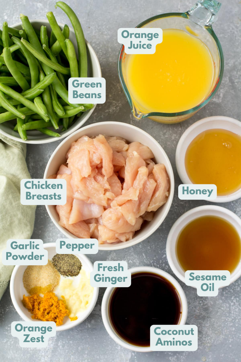 Ingredients for Healthy Orange Chicken in small white bowls on a grey background. Green beans, chicken, orange juice, honey, toasted sesame oil, coconut aminos and spices