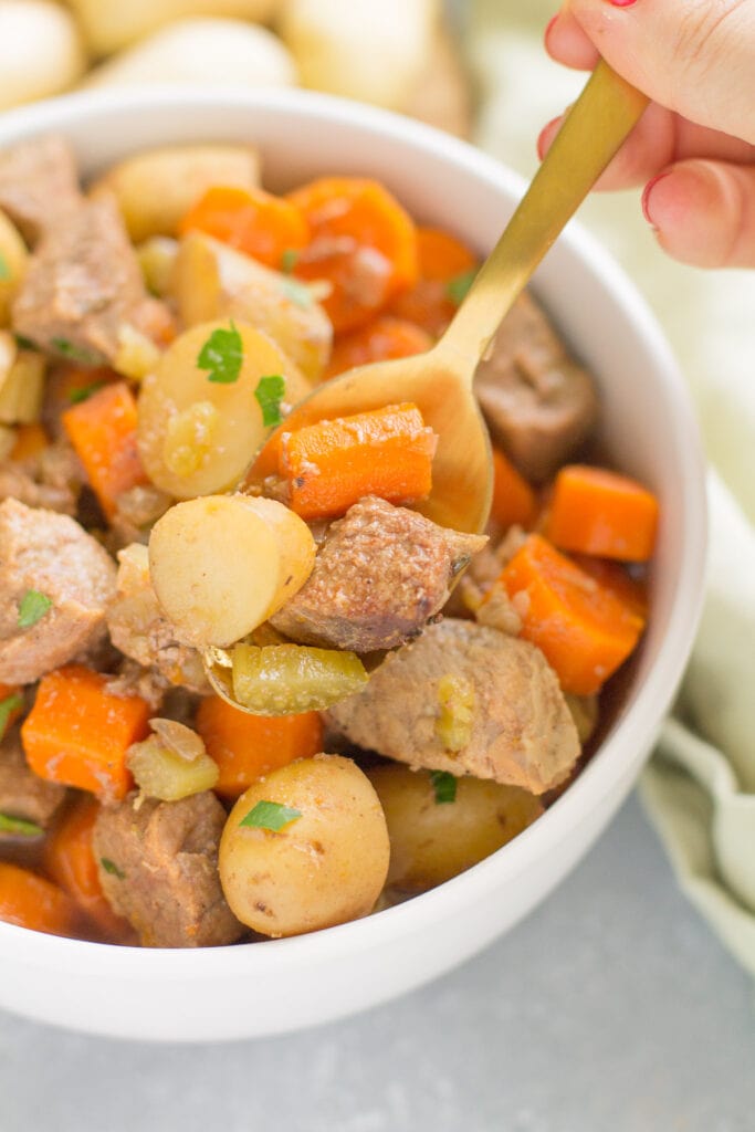 Paleo Beef stew in a white bowl with a green linen in the background. A gold spoon scooping a bite out of it.