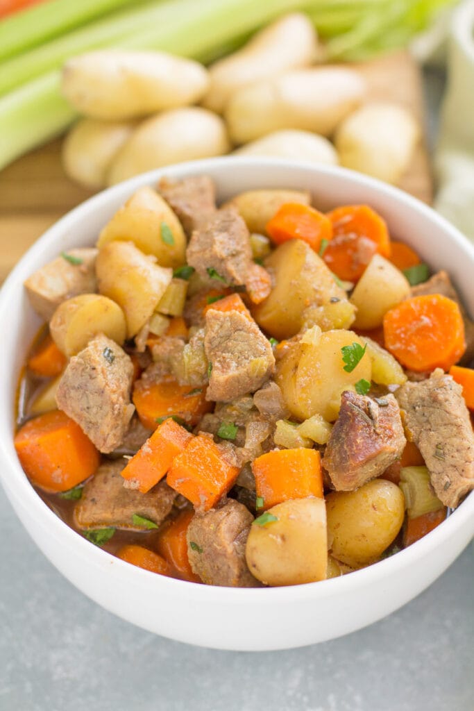 A close up of healthy beef stew with a cutting board in the background that has carrots, potatoes and celery in the background