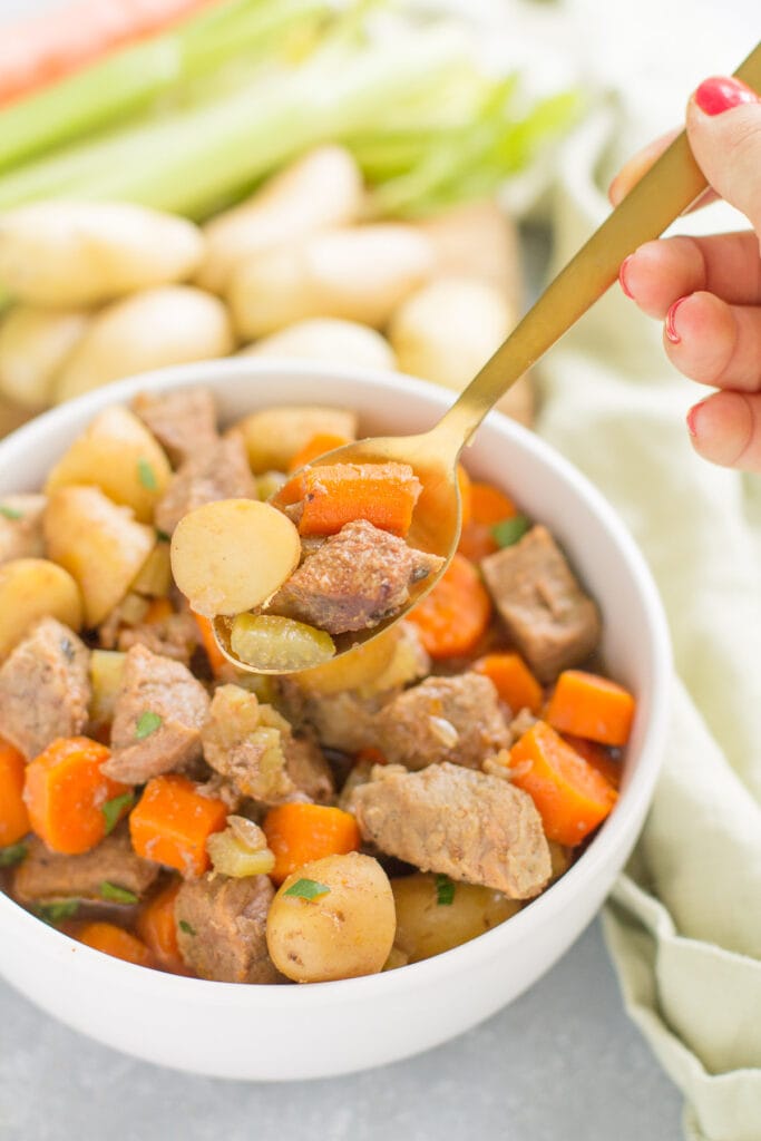 A gold spoon taking a bite out of healthy paleo beef stew that's in a white bowl with a green linen in the background
