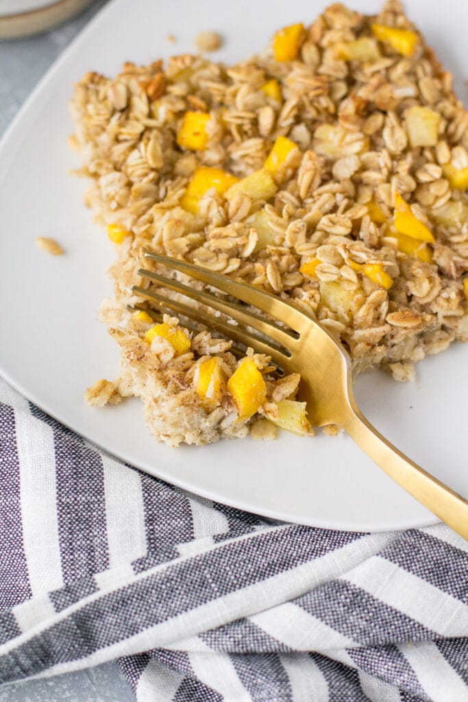 A close up of mango pineapple oatmeal on a white plate with a gold fork taking a bite out of it.
