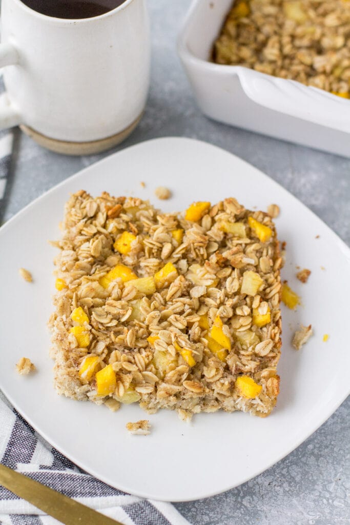 A close up of pineapple baked oatmeal on a white plate on a grey background with coffee in the background.