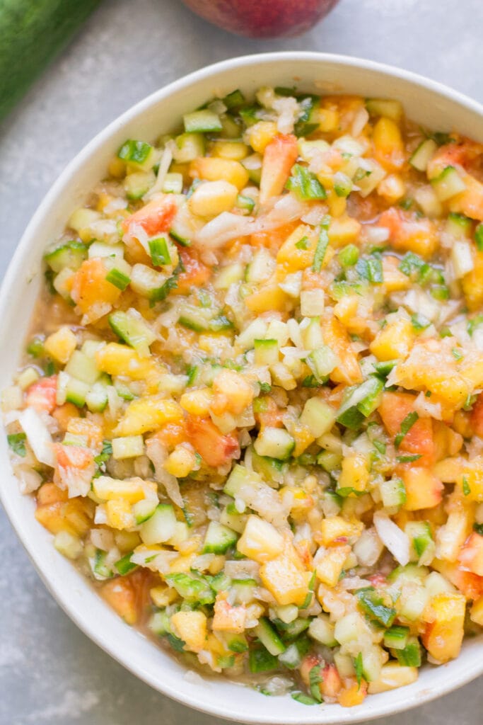Peach salsa in a white bowl on a grey background with cucumber and peaches in the background