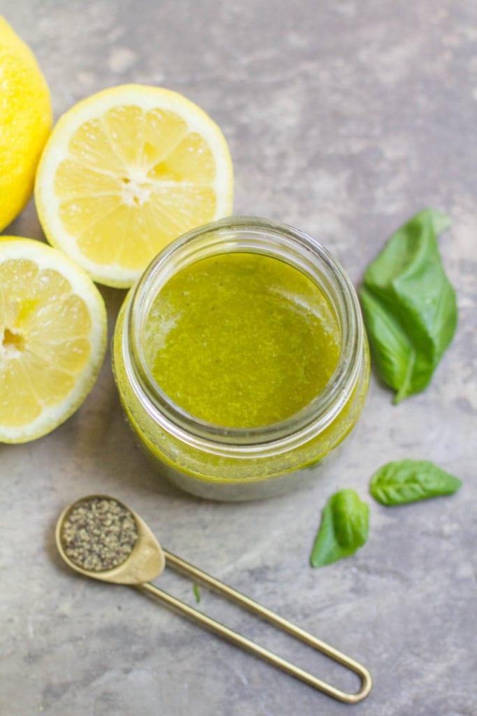 Overhead view of lemon basil vinaigrette in a small glass jar with fresh herbs and lemon halves scattered around it.