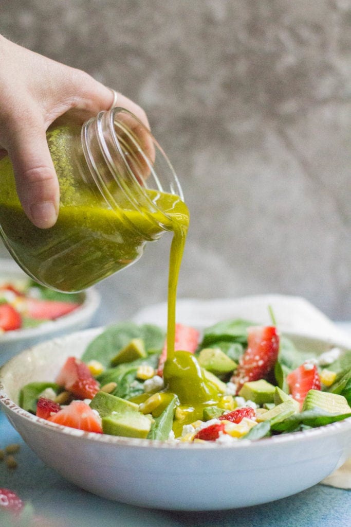 Hand holding a glass jar with lemon vinaigrette, pouring it over a bowl full of salad.