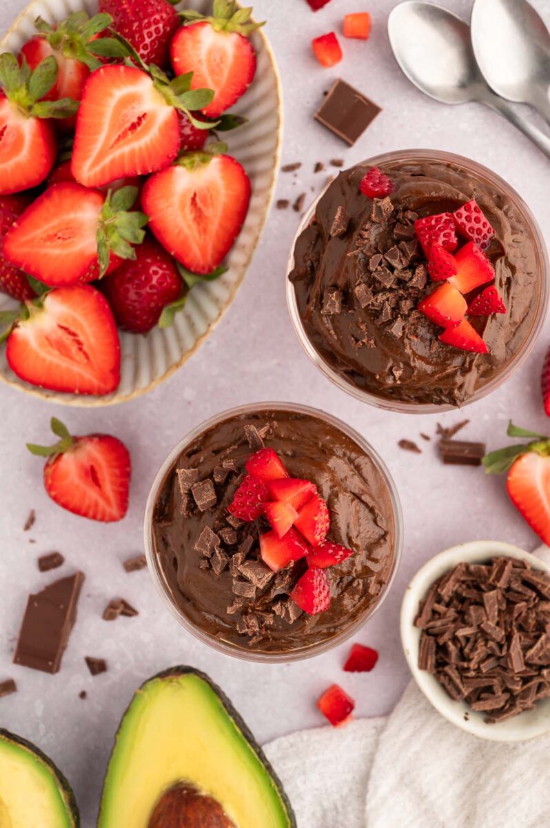 An overhead shot of two dishes of healthy chocolate pudding topped with chopped strawberries and a bowl of strawberries and shaved chocolate on the side.