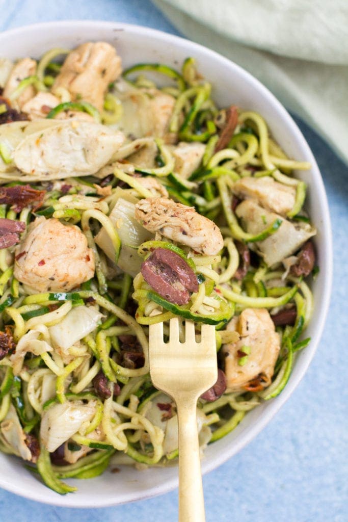 Whole30 Greek Chicken Zucchini Noodles in a white bowl with a gold fork taking out a bite on a blue background