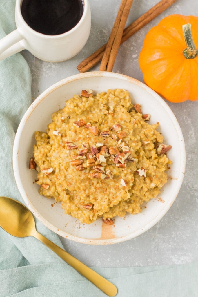 An overhead shot of pumpkin slow cooker steel cut oats on a grey background with a cup of coffee, cinnamon sticks and a small pumpkin in the background