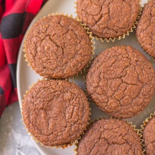 A white plate with healthy gingerbread muffins stacked on it. A red checked linen, cinnamon sticks + ornaments are scattered in the background
