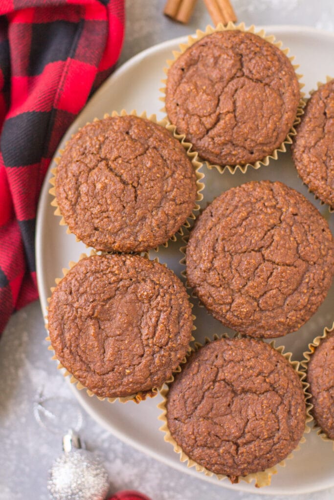 A white plate with gingerbread muffins stacked on it. A red checked linen, cinnamon sticks + ornaments are scattered in the background