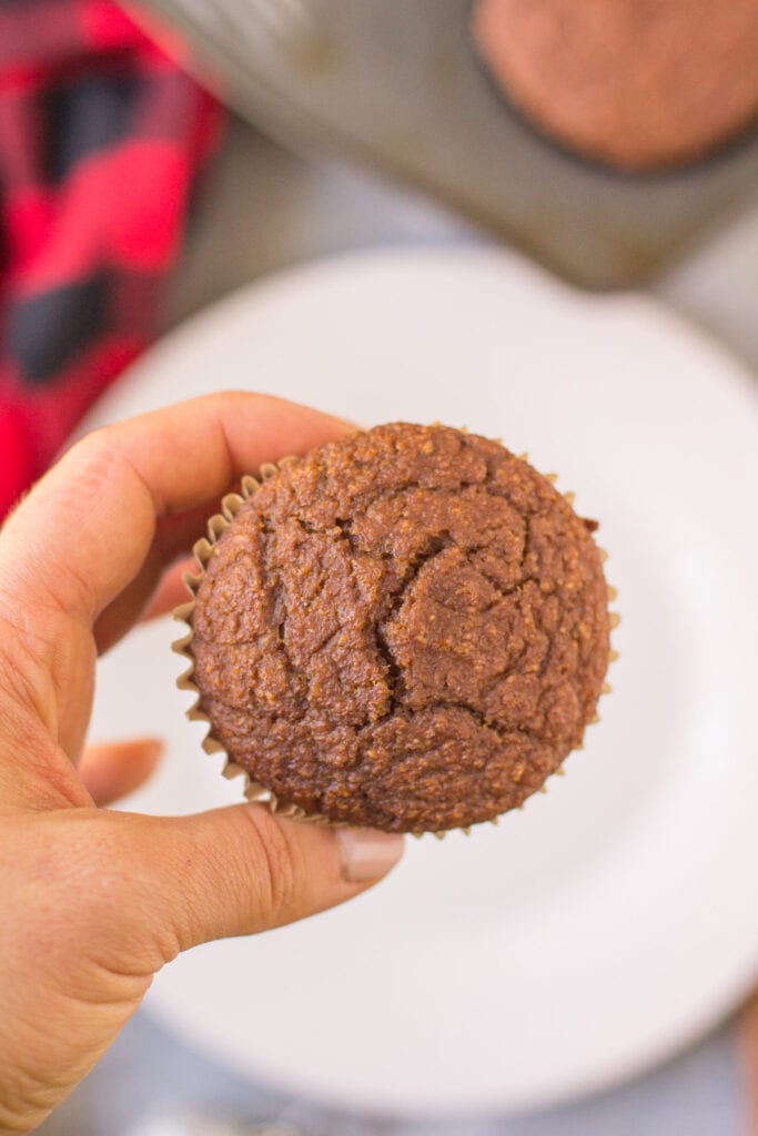 A hand holding a gingerbread muffin with a red linen and a muffin tin in the background