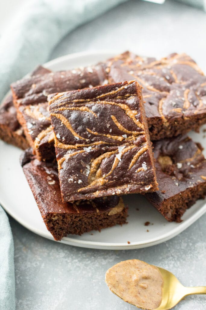 A plate of almond butter brownies with a scoop of almond butter off to the side and a blue linen in the background.