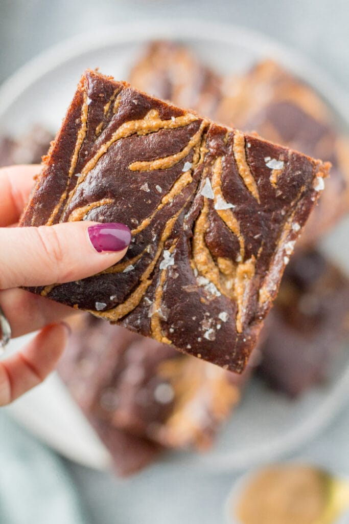 A hand holding a salted almond butter brownie up to the camera with a plate of brownies in the background
