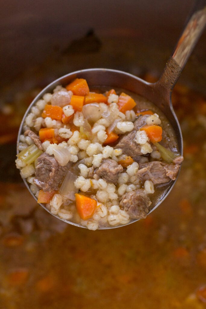 A ladle holding up a scoop beef barley soup with chunks of beef stew meat