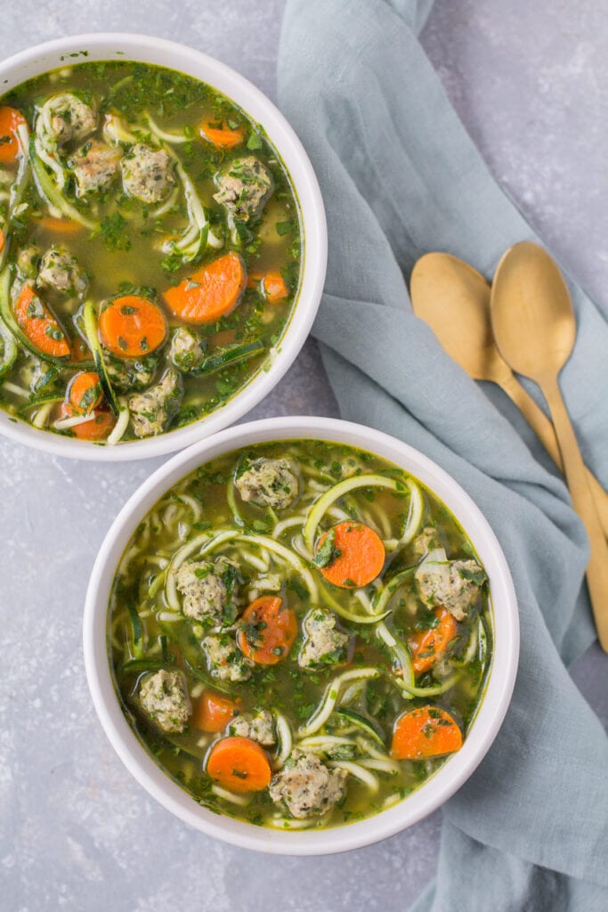 Two bowls of low calorie Italian Wedding Soup on a grey background. a blue linen and gold spoons sit off to the side.