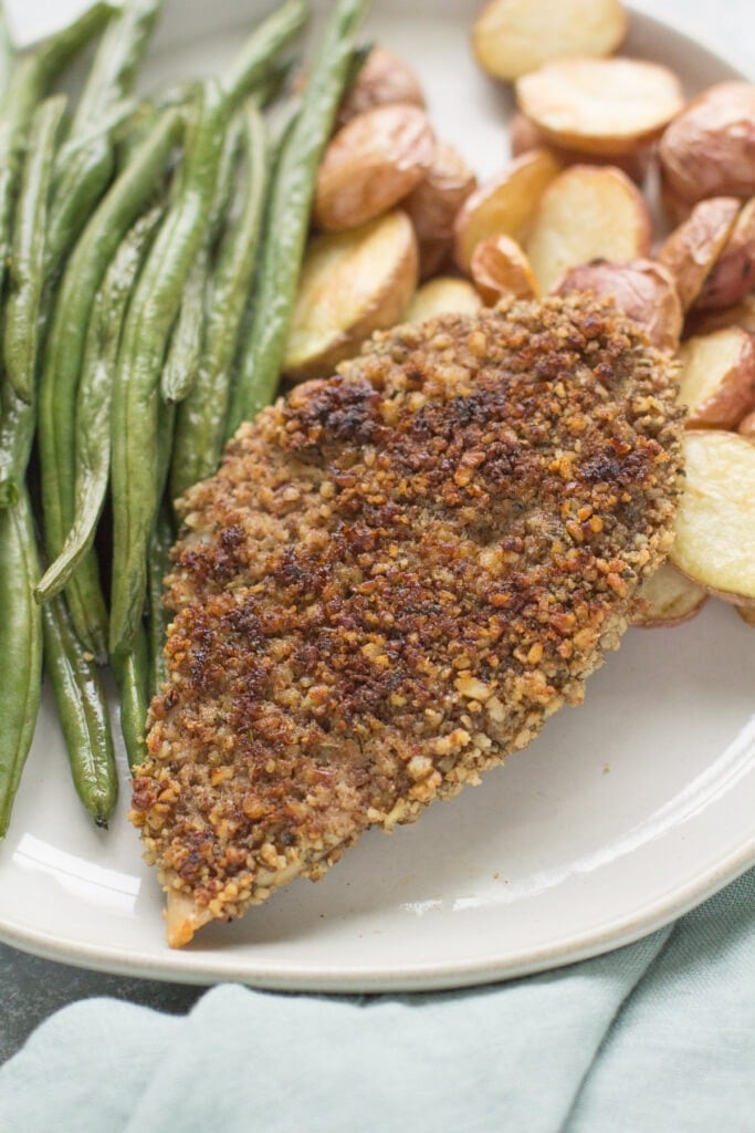 A close up of pecan chicken cutlets on a white plate with roasted potatoes and green beans off to the side