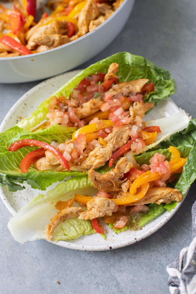 Whole30 Chicken Fajitas in lettuce wraps on a white plate on a grey background with a pan of fajitas in the background