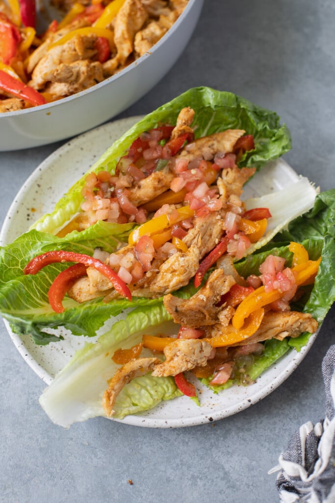 Healthy Chicken Fajitas in lettuce wraps on a white plate on a grey background with a pan of fajitas in the background