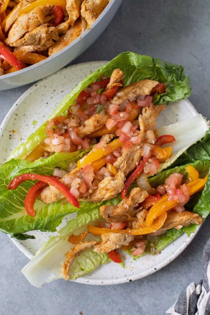 Healthy Chicken Fajitas in lettuce wraps on a white plate on a grey background with a pan of fajitas in the background