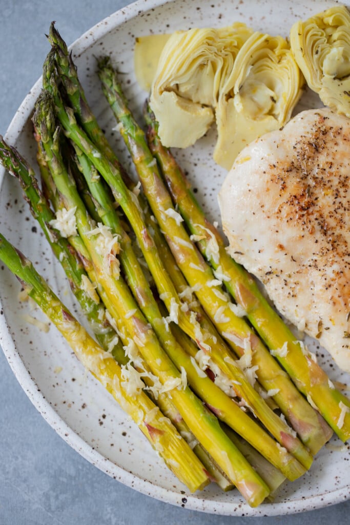 Healthy Lemon Parmesan Roasted Asparagus on a speckled plate with chicken and artichoke hearts on a grey background