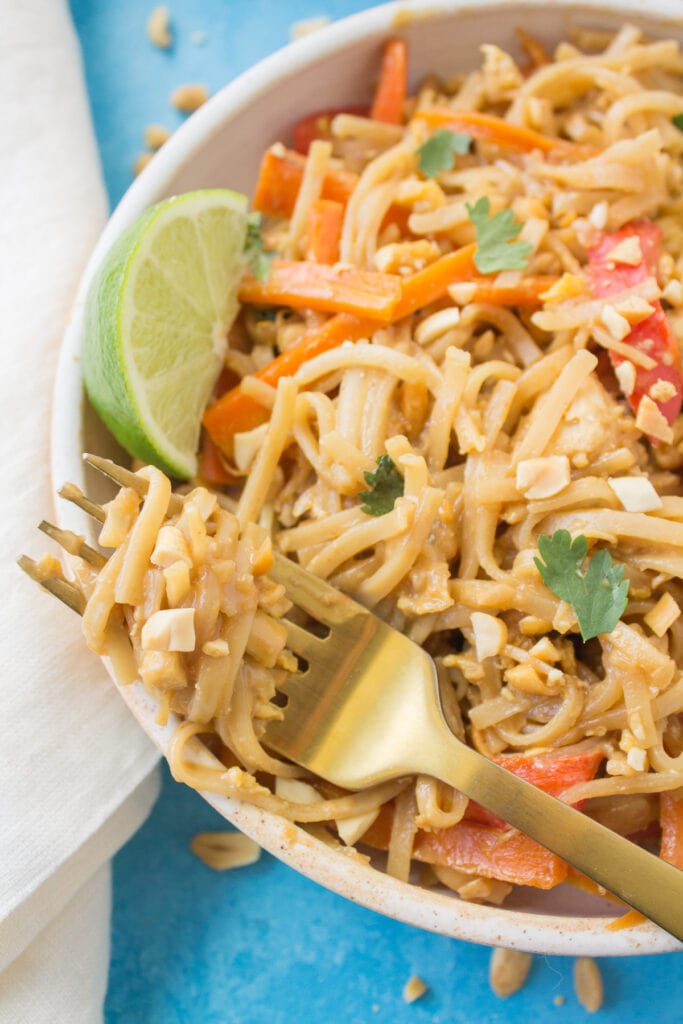 A white bowl filled with Healthier Chicken Pad Thai. A gold fork is off to the side with a bite of noodles swirled on to it with a white linen in the background