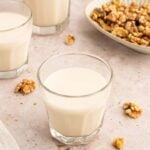 Three clear glasses of homemade walnut milk with walnuts spread around and a bowl of walnuts in the background.