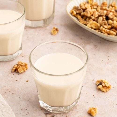 Three clear glasses of homemade walnut milk with walnuts spread around and a bowl of walnuts in the background.