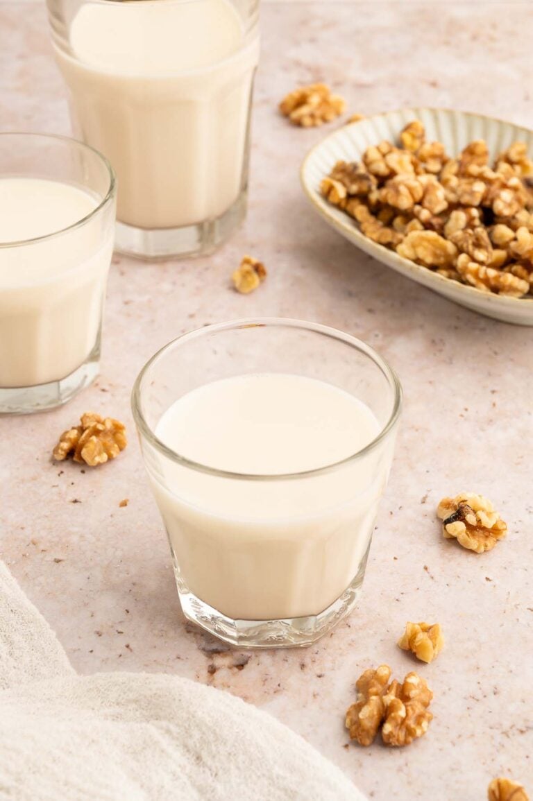Three clear glasses of homemade walnut milk with walnuts spread around and a bowl of walnuts in the background.