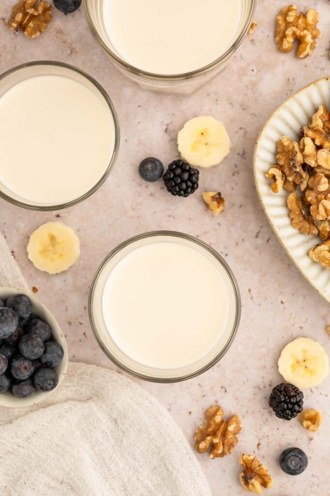 Three glasses of homemade walnut milk with blueberries, walnuts, and sliced bananas on the counter.