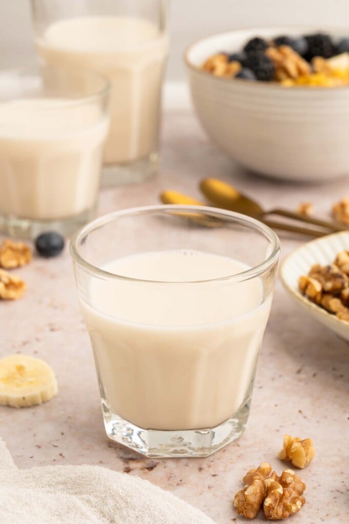 A clear glass of walnut milk with more glasses and a bowl of cereal in the background.