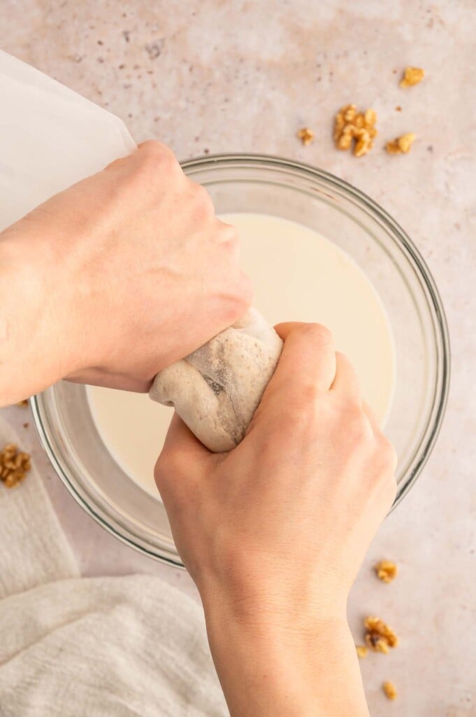 Two hands squeezing a nut milk bag with a bowl of walnut milk in the background.