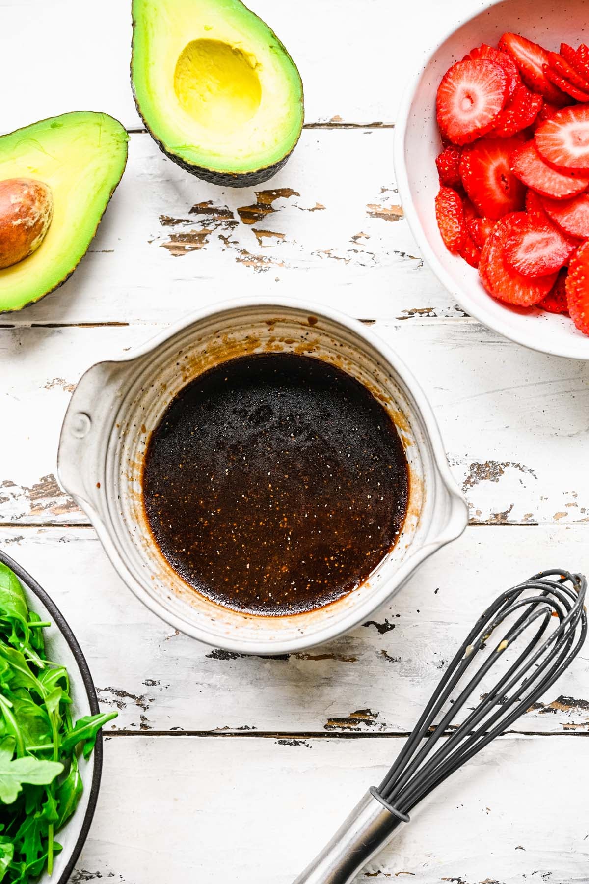 A bowl of vinaigrette for strawberry arugula salad with strawberries, avocado, arugula on the side