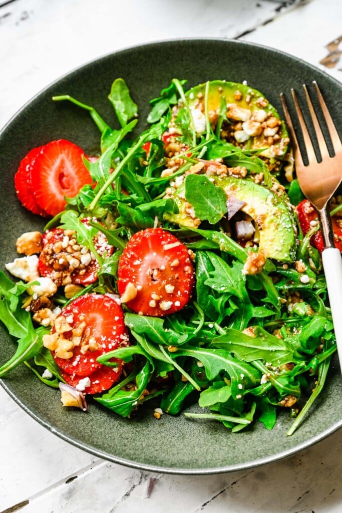 A close up of a grey plate with strawberry arugula salad with a fork on it on a white background