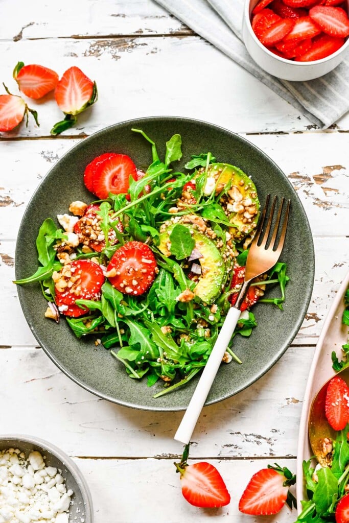 An overhead of a grey plate with strawberry arugula salad with a fork on it on a white background