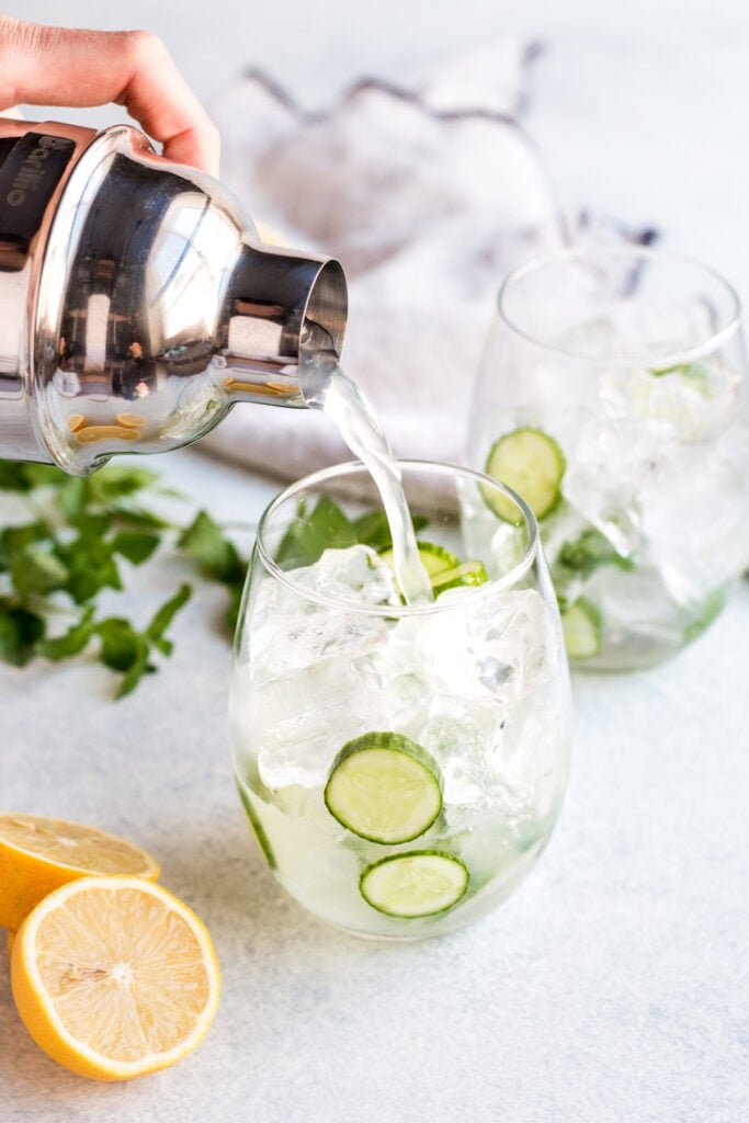 A cocktail shaker pouring a cucumber vodka drink into a glass with ice, lemons and cucumbers