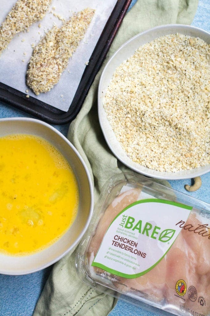 Two bowls, a package of chicken tenders and a baking sheet with battered chicken. In one bowl is whisked eggs, in the other bowl is coconut cashew crust.