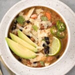Crockpot Chicken Quinoa Soup in a white bowl garnished with avocado on a white plate with a grey background
