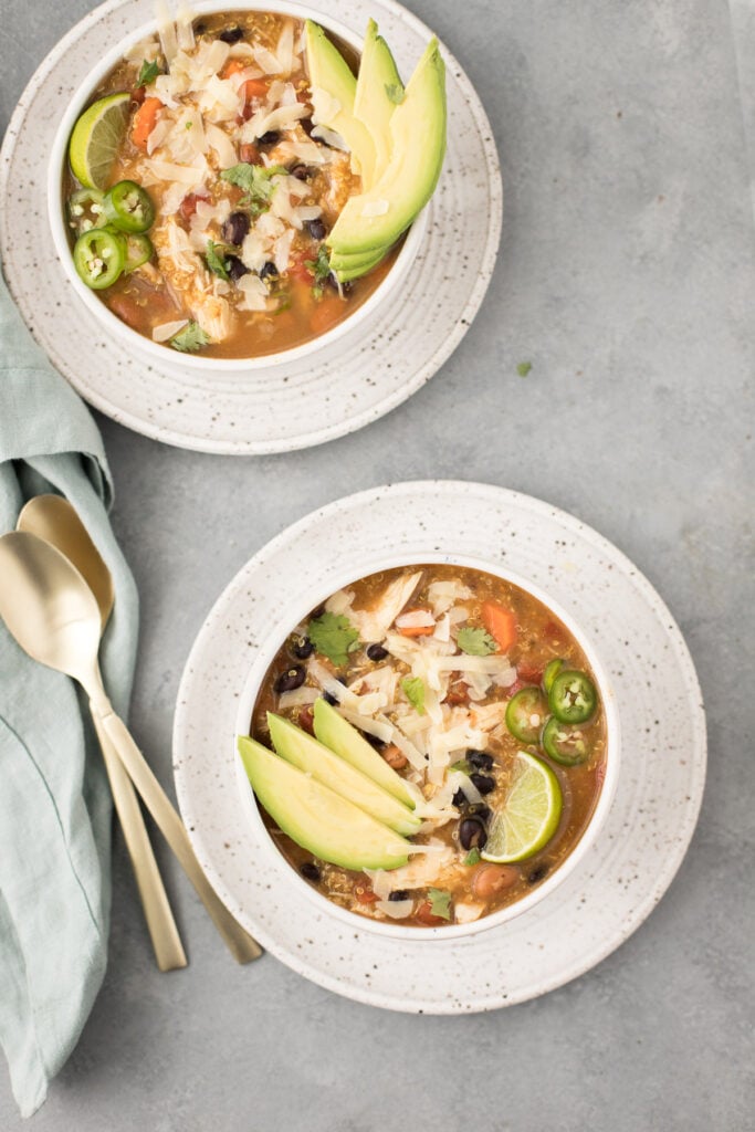 Two bowls of Chicken Quinoa Soup on a grey background with spoons and a blue linen off to the side.