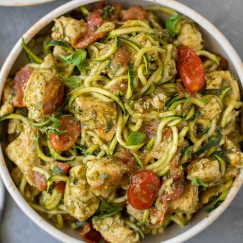 Whole30 Pesto Chicken Zucchini Noodles in a white bowl with tomatoes and basil scattered around.