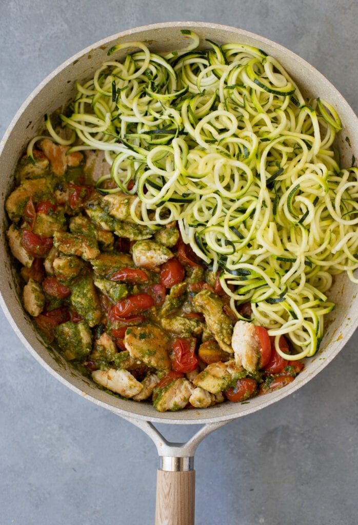 Pesto chicken zoodles in a pan before being tossed together. zoodles are off to the side and pesto/chicken/tomato mixture is on the other side