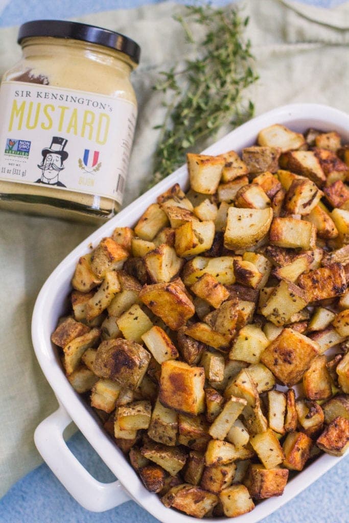 Crispy Dijon Roasted Potatoes in a white bowl on a blue background with Sir Kensington's mustard and thyme leaves off to the side