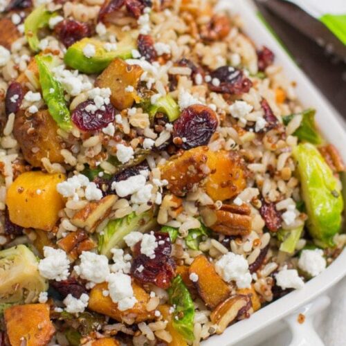 Harvest Quinoa and Brown Rice Salad in a white bowl with a blue background. The salad is filled with brown rice, quinoa, cranberries, btuternut squash and brussels sprouts