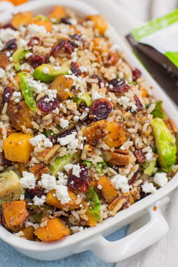 Harvest Quinoa and Brown Rice Salad in a white bowl with a blue background. The salad is filled with brown rice, quinoa, cranberries, btuternut squash and brussels sprouts