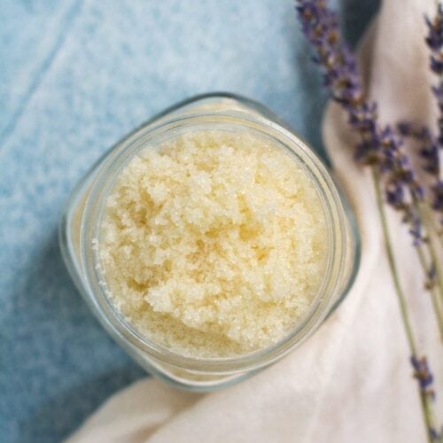 Easy DIY Sugar Scrub in a small glass jar on a blue background. A white linen and lavender off to the side on the right.