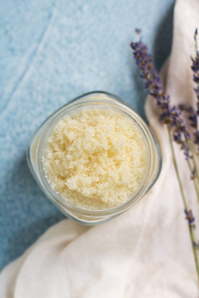 Easy DIY Sugar Scrub in a small glass jar on a blue background. A white linen and lavender off to the side on the right.