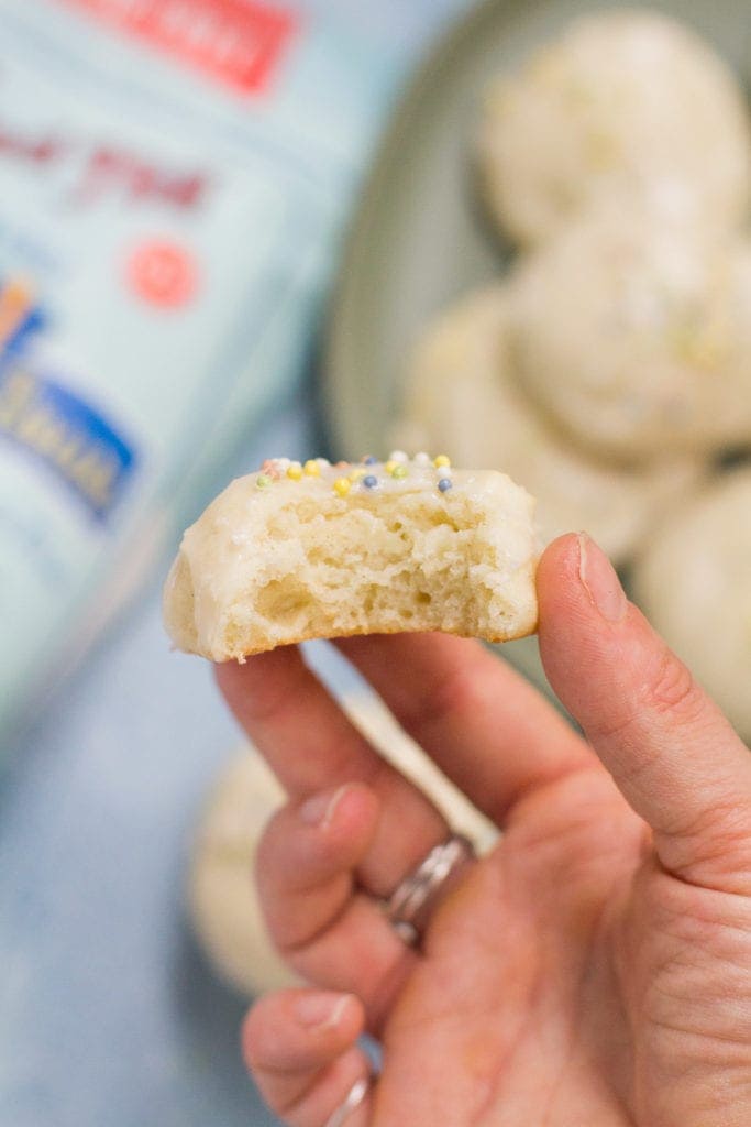 A hand holding up Healthy Anginetti Cookies with a bite taken out of one. Bob's Red Mill Gluten Free Flour off to the left corner in the background.