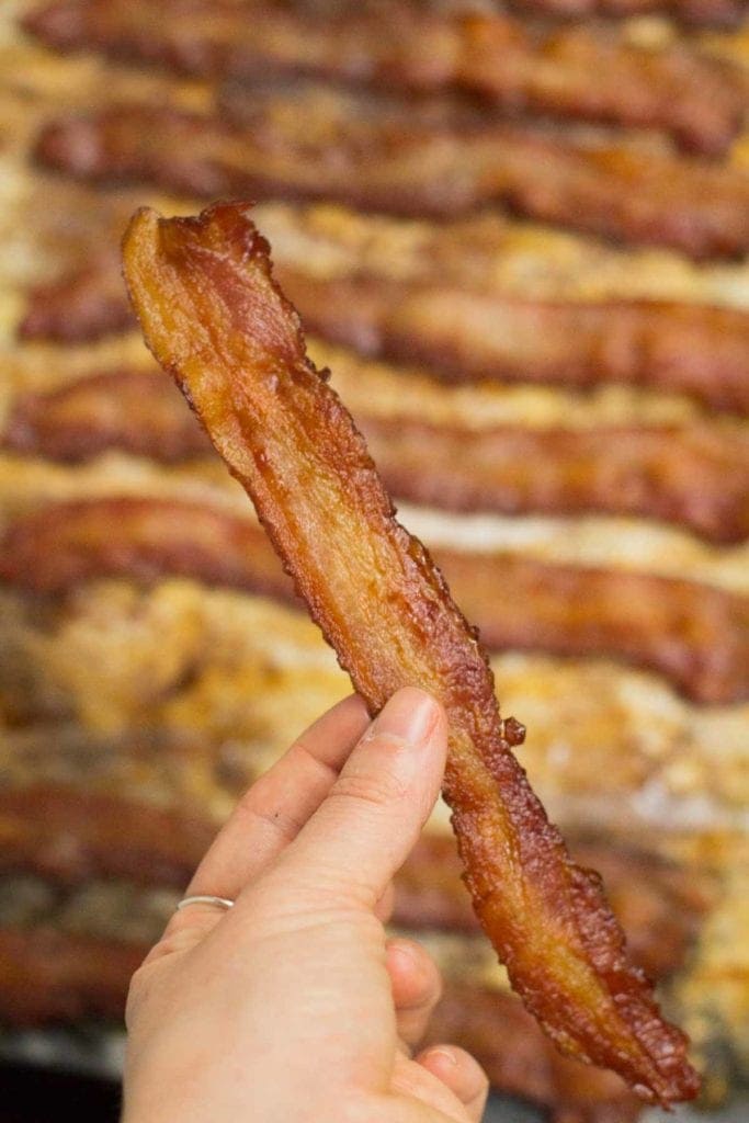 a piece of crispy bacon being held up by a hand with a sheet of bacon in the background.