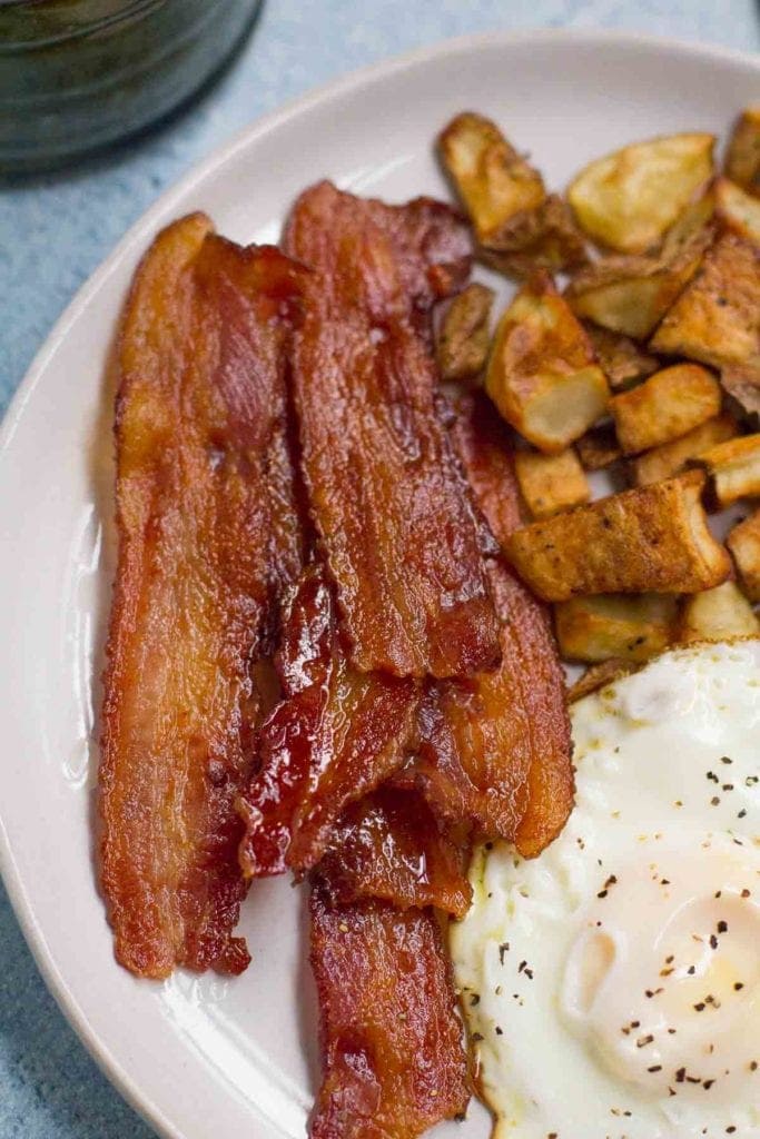 a white plate with oven roasted crispy bacon, a fried egg and crispy potatoes with a gold fork off to the side.